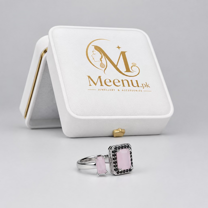A silver ring with a rectangular light pink stone and a black diamond-like border, displayed in the foreground with its branded white Meenu jewelry box in the background against a dark gray backdrop.