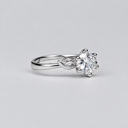 A silver ring with a clear gemstone is displayed in the foreground on a light surface, with a white jewelry box branded 'Meenu' in the blurred background.