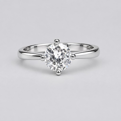 A silver solitaire ring with a clear round gemstone is displayed on a light surface, with a blurred white jewelry box featuring an orange 'M' logo in the background.