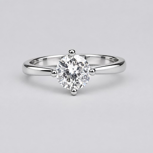 A silver solitaire ring with a clear round gemstone is displayed on a light surface, with a blurred white jewelry box featuring an orange 'M' logo in the background.