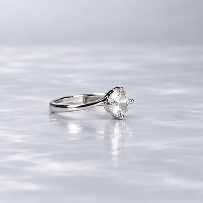 A silver ring with a clear gemstone is displayed on a light lavender surface, with its branded white presentation box in the background against a dark gray backdrop.