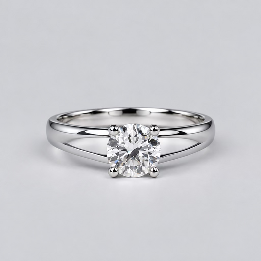 A silver ring with a clear central gemstone is displayed on a light surface, with a white Meenu jewelry box in the blurred background.