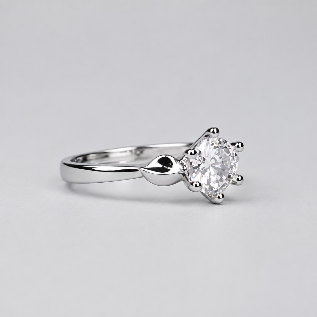 A silver solitaire engagement ring with a clear round-cut gemstone, displayed on a light lavender surface with a white jewelry box branded 'Meenu Jewels' in the blurred background.
