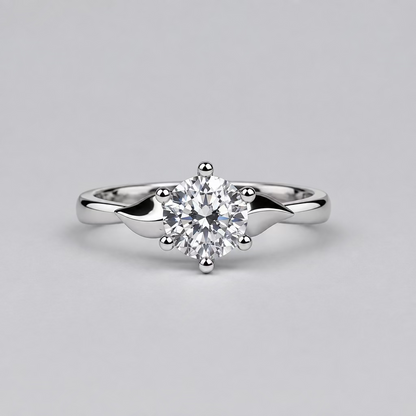 A silver engagement ring with a clear round-cut center stone is displayed on a light surface, with a white Meenu jewelry box in the blurred background.