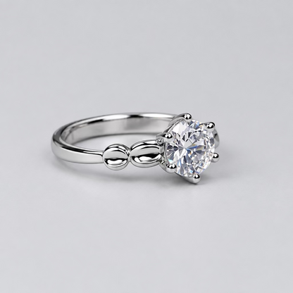 A silver-toned solitaire ring with a clear round gemstone, displayed on a light surface with a blurred white jewelry box featuring an 'M' logo in the background.