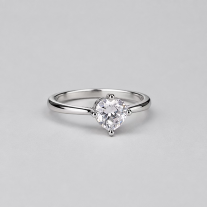 A silver solitaire engagement ring with a round-cut clear gemstone, displayed on a light lavender surface with a blurred white jewelry box featuring an orange 'M' logo in the background.