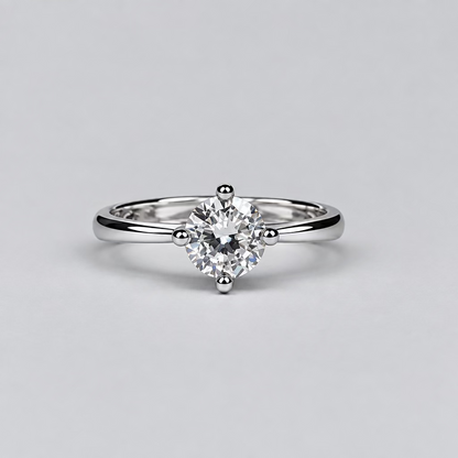 A silver-toned ring with a round clear gemstone is displayed on a light surface, with an open white jewelry box featuring the Meenu jewelry brand in the blurred background.
