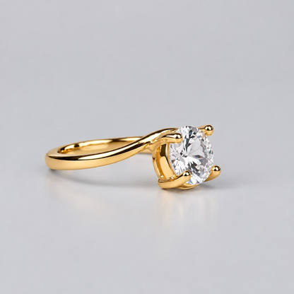 A gold ring with a clear gemstone is displayed on a light surface, with a white jewelry box featuring the 'Meenu Jewels' logo in the blurred background.