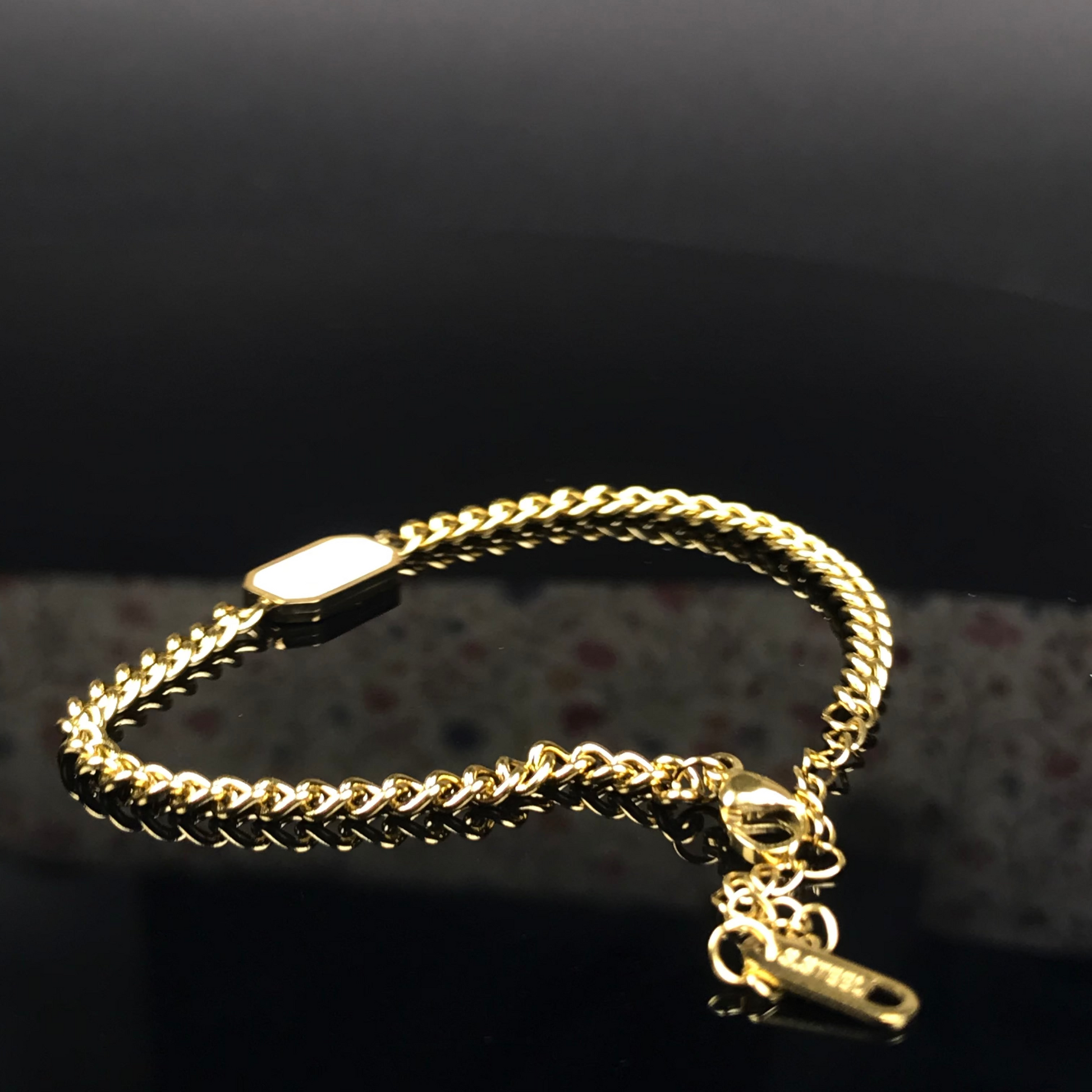 A gold chain bracelet with a lobster clasp and a small hanging tag, photographed on a dark reflective surface.