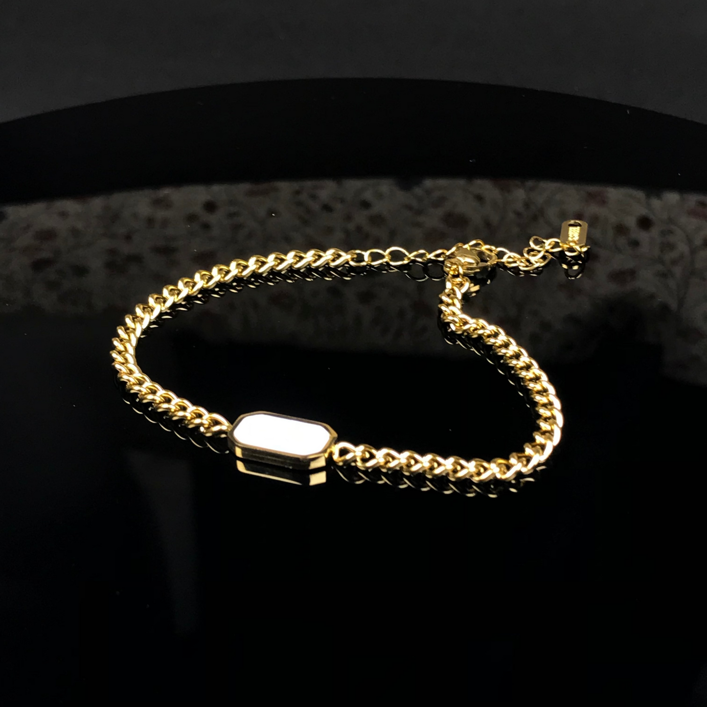 A gold-toned chain bracelet with a polished rectangular white inlay, displayed on a reflective black surface.