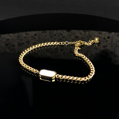 A gold-toned chain bracelet with a polished rectangular white inlay, displayed on a reflective black surface.