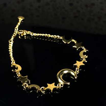 A gold-toned chain bracelet featuring dangling crescent moon and star charms, displayed on a reflective black surface.
