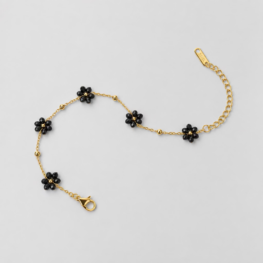A delicate gold chain bracelet featuring five small black flower charms, laid out on a light pink background.