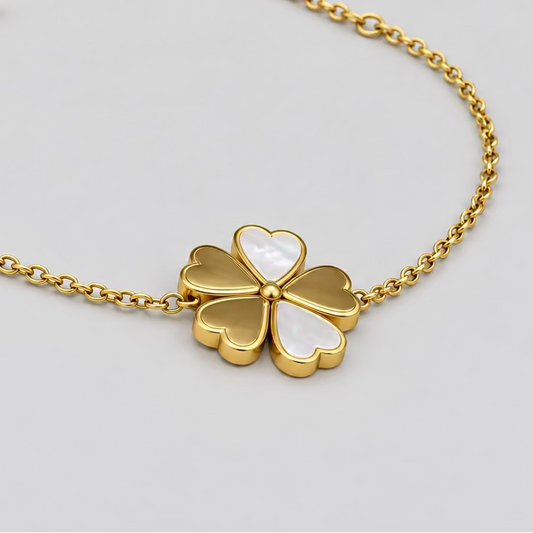 A delicate gold chain bracelet featuring a four-leaf clover charm with a white inlay, resting on a light pink surface with a dark background.
