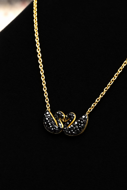 A gold chain necklace with a pendant featuring two black swan figures adorned with small clear stones, displayed on a black velvet surface.