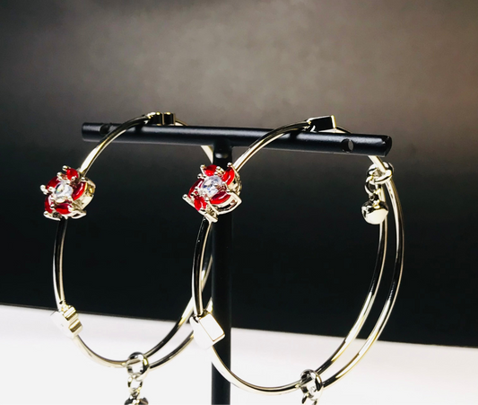 Gold hoop earrings with red and clear crystal floral accents, displayed on a black stand against a dark gray background.