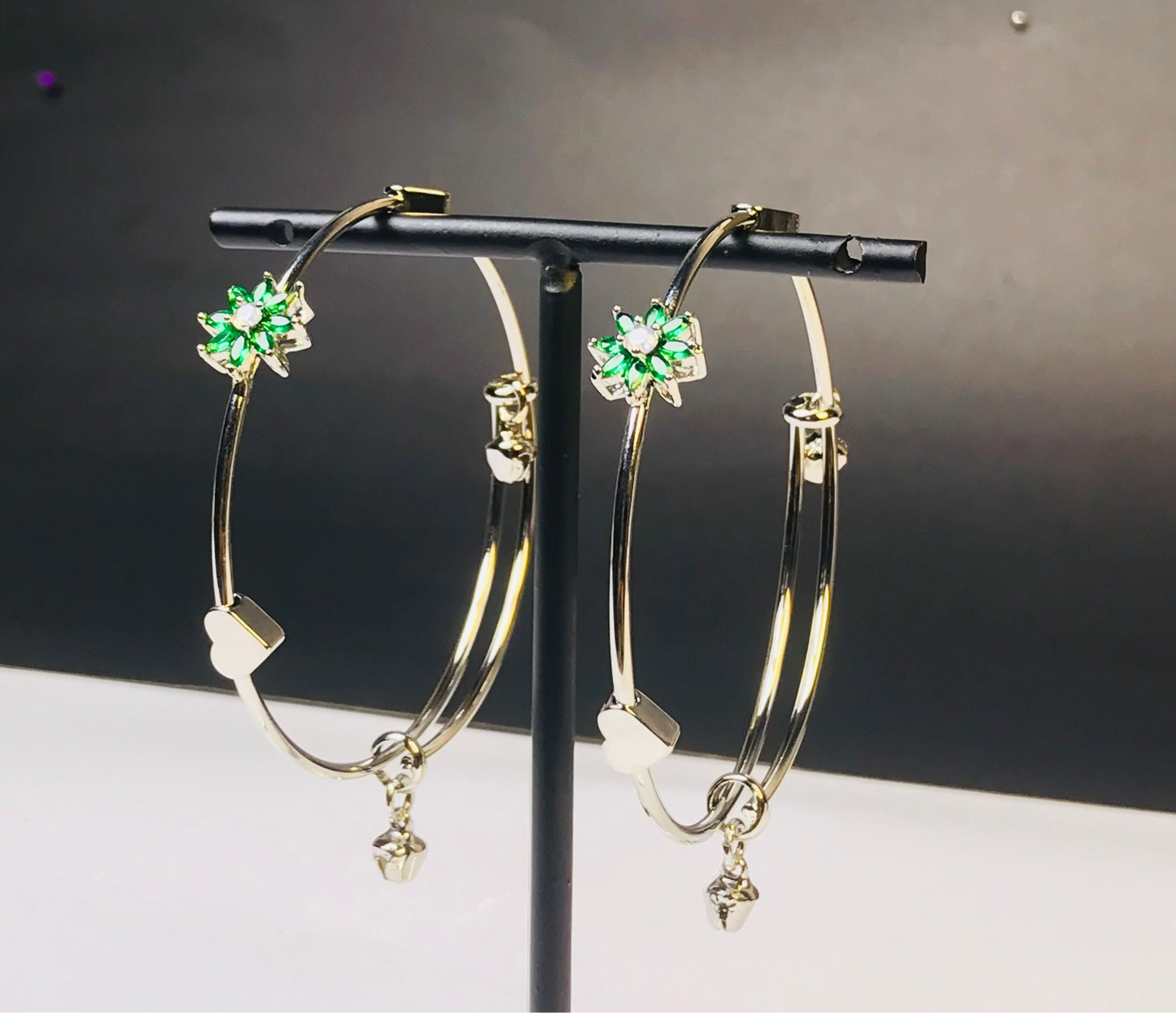 Gold hoop earrings with green four-leaf clover charms and dangling heart charms, displayed on a black stand.