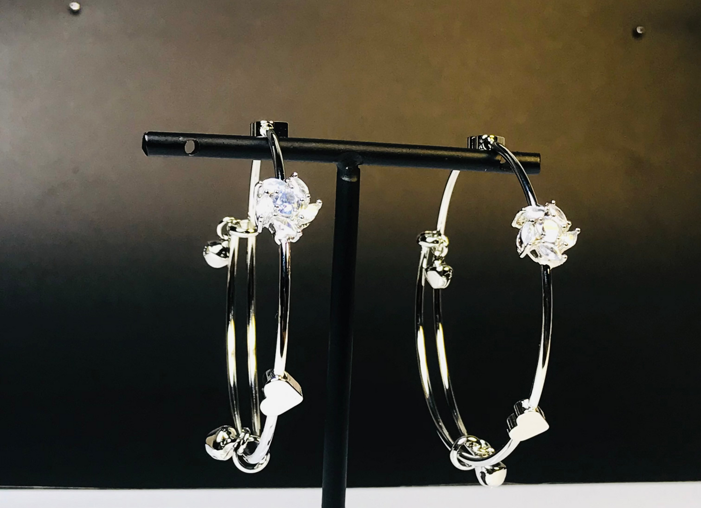 A pair of silver hoop earrings with clear faceted gemstones and small dangling charms, displayed on a black stand against a dark brown background.