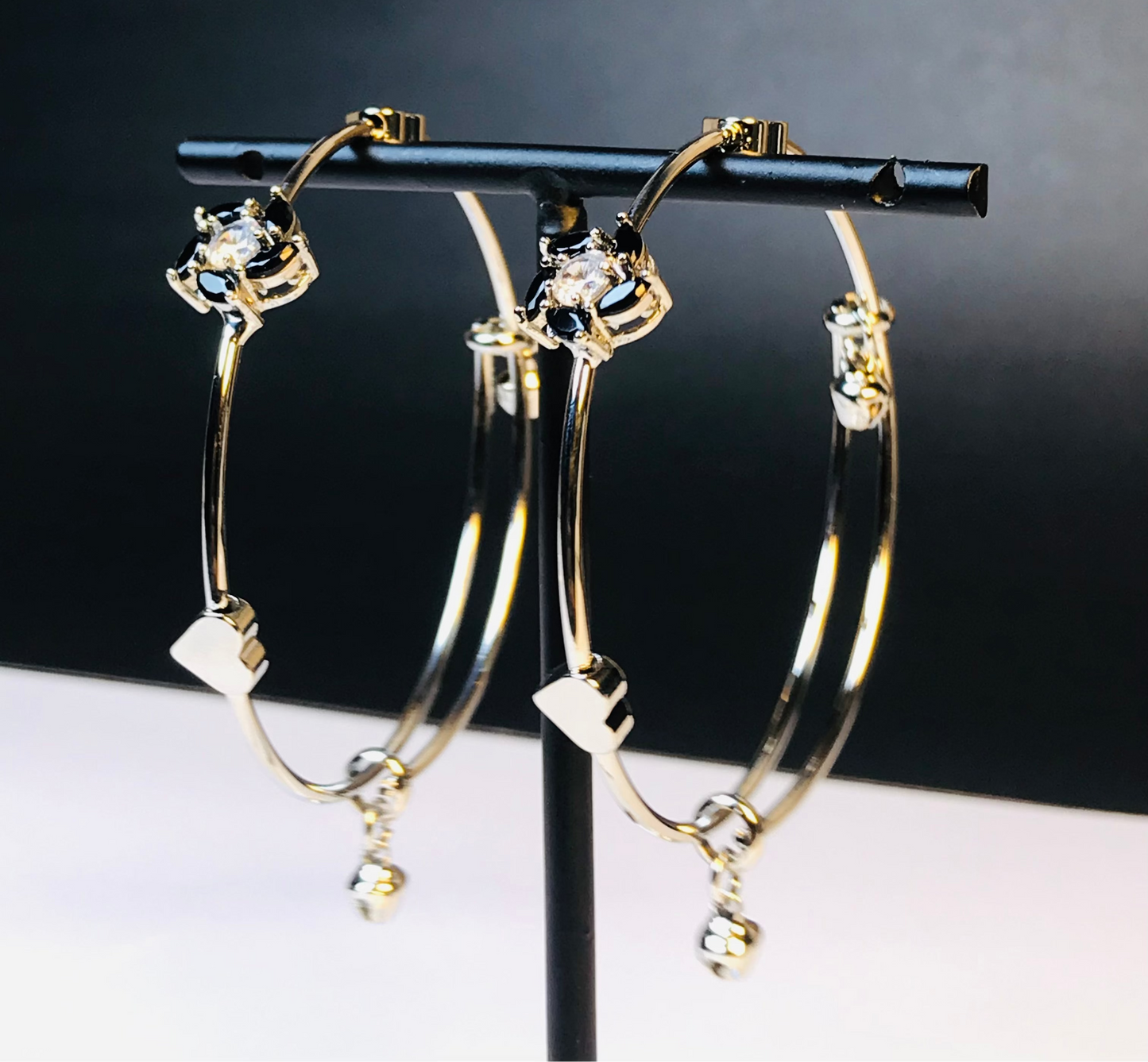Gold hoop earrings with clear rhinestone details and small dangling charms, displayed on a black jewelry stand against a dark background.