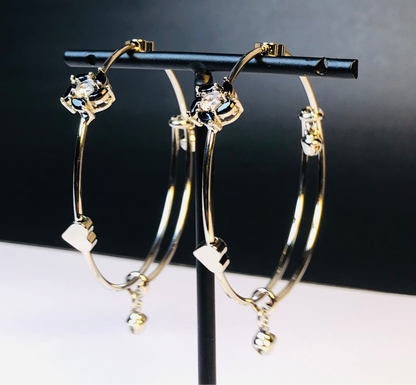 Gold hoop earrings with clear rhinestone details and small dangling charms, displayed on a black jewelry stand against a dark background.
