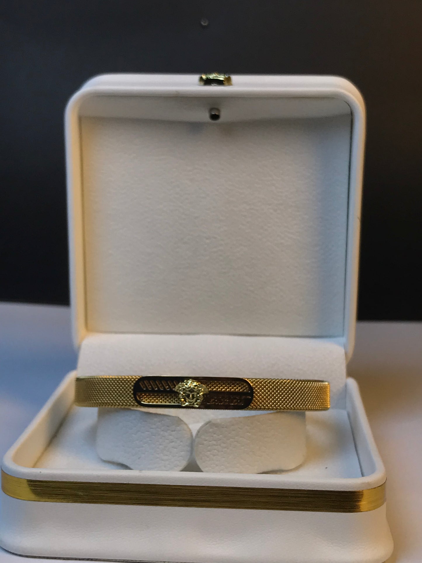 A gold Versace bracelet with a textured mesh band and a central Medusa emblem, displayed inside an open white velvet jewelry box.
