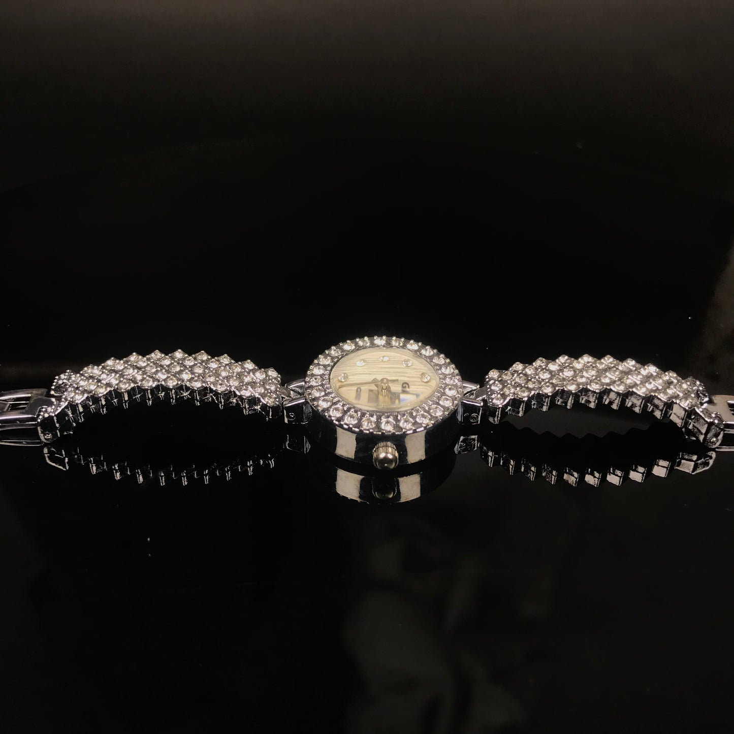 A silver-tone watch with a round dial and a wide bracelet covered in clear rhinestones, displayed on a glossy black surface against a dark background.