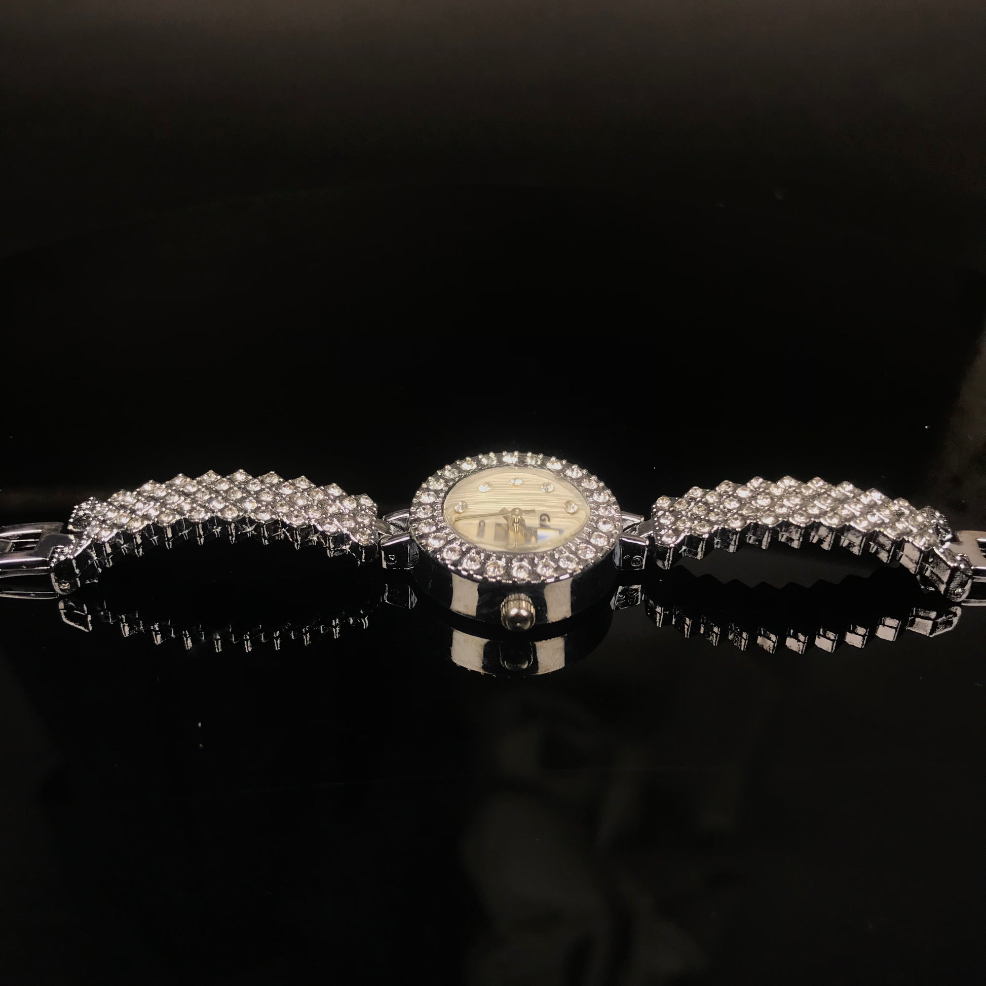 A silver-tone watch with a round dial and a wide bracelet covered in clear rhinestones, displayed on a glossy black surface against a dark background.