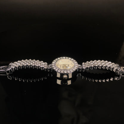 A silver-tone watch with a round dial and a wide bracelet covered in clear rhinestones, displayed on a glossy black surface against a dark background.
