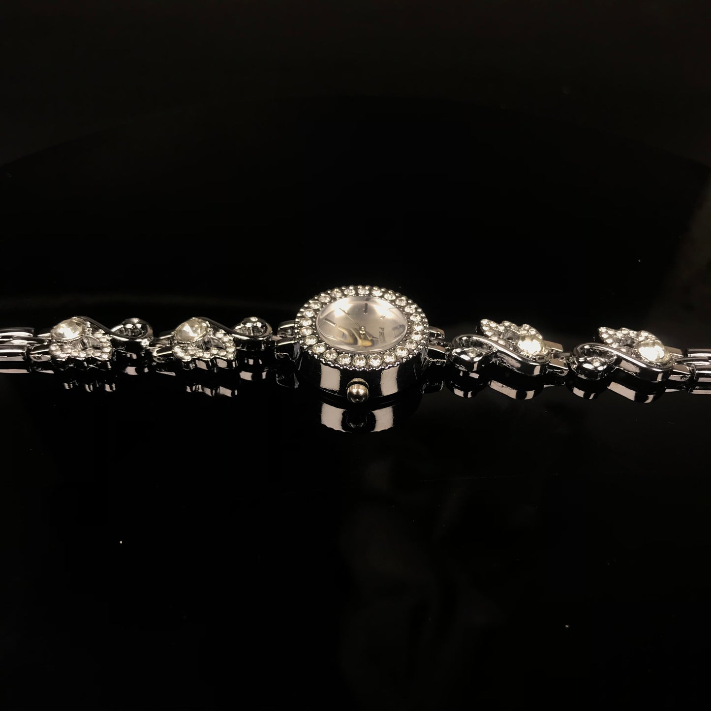 A silver-toned bracelet watch with a round crystal-studded bezel, a light dial, and matching crystal-studded links on a black reflective background.