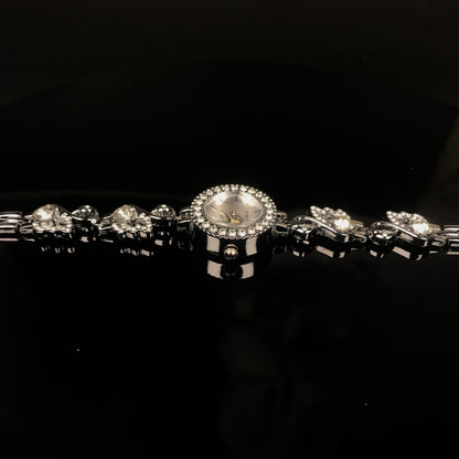 A silver-toned bracelet watch with a round crystal-studded bezel, a light dial, and matching crystal-studded links on a black reflective background.