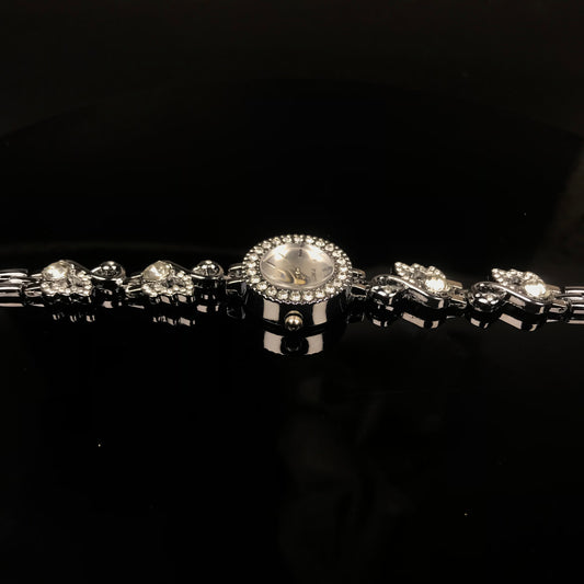 A silver-toned bracelet watch with a round crystal-studded bezel, a light dial, and matching crystal-studded links on a black reflective background.