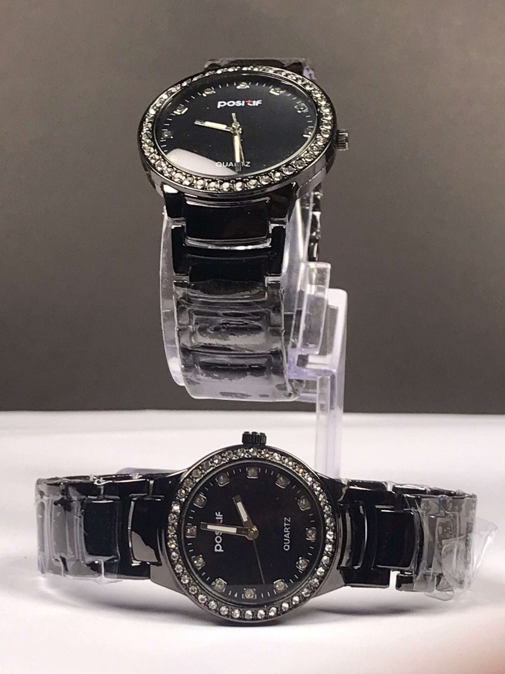 Two black couples quartz wristwatches with rhinestone-studded bezels and black crocodile-embossed straps, displayed on clear plastic stands against a gray and white background.