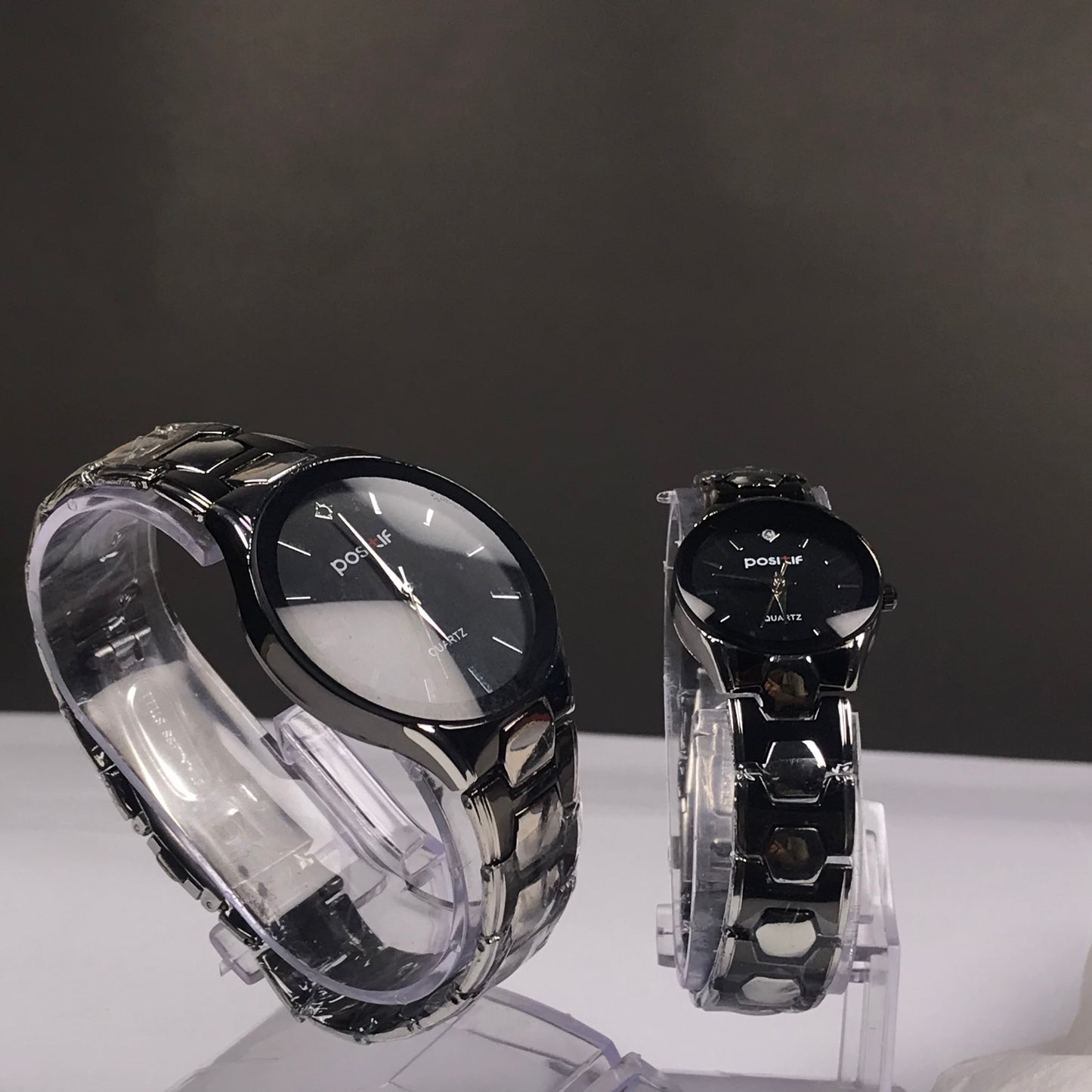 Two black analog wristwatches with metallic links, displayed on clear acrylic stands against a dark gray background.