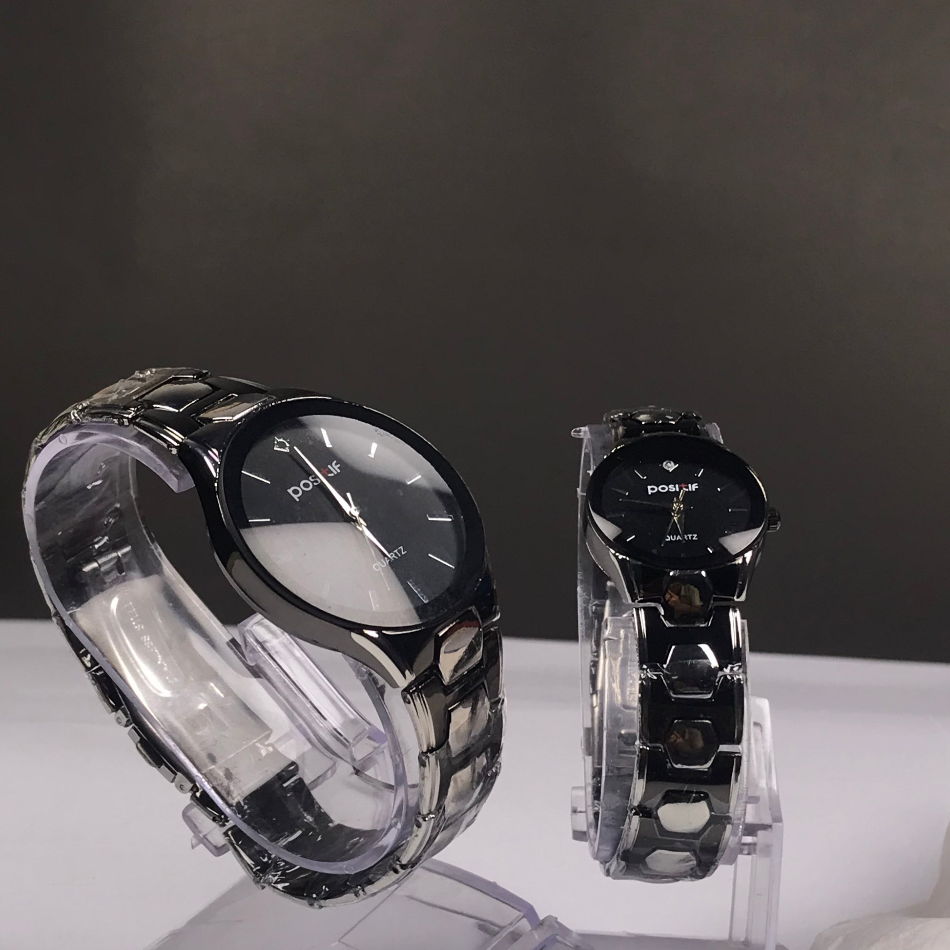 Two black analog wristwatches with metallic links, displayed on clear acrylic stands against a dark gray background.