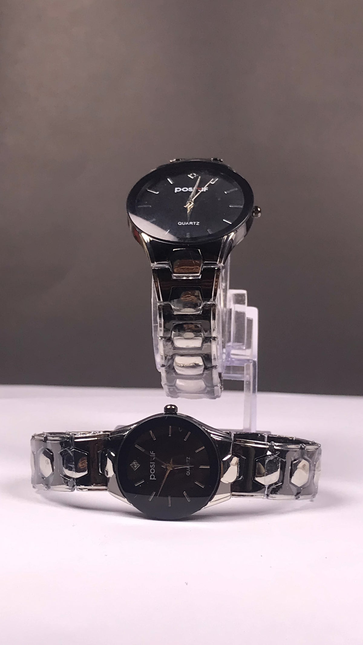 Two black analog wristwatches with silver metal link bracelets displayed on clear stands against a gray studio background.