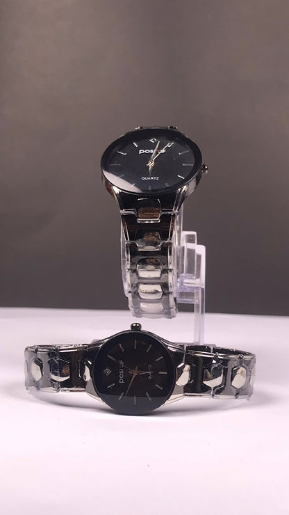 Two black analog wristwatches with silver metal link bracelets displayed on clear stands against a gray studio background.