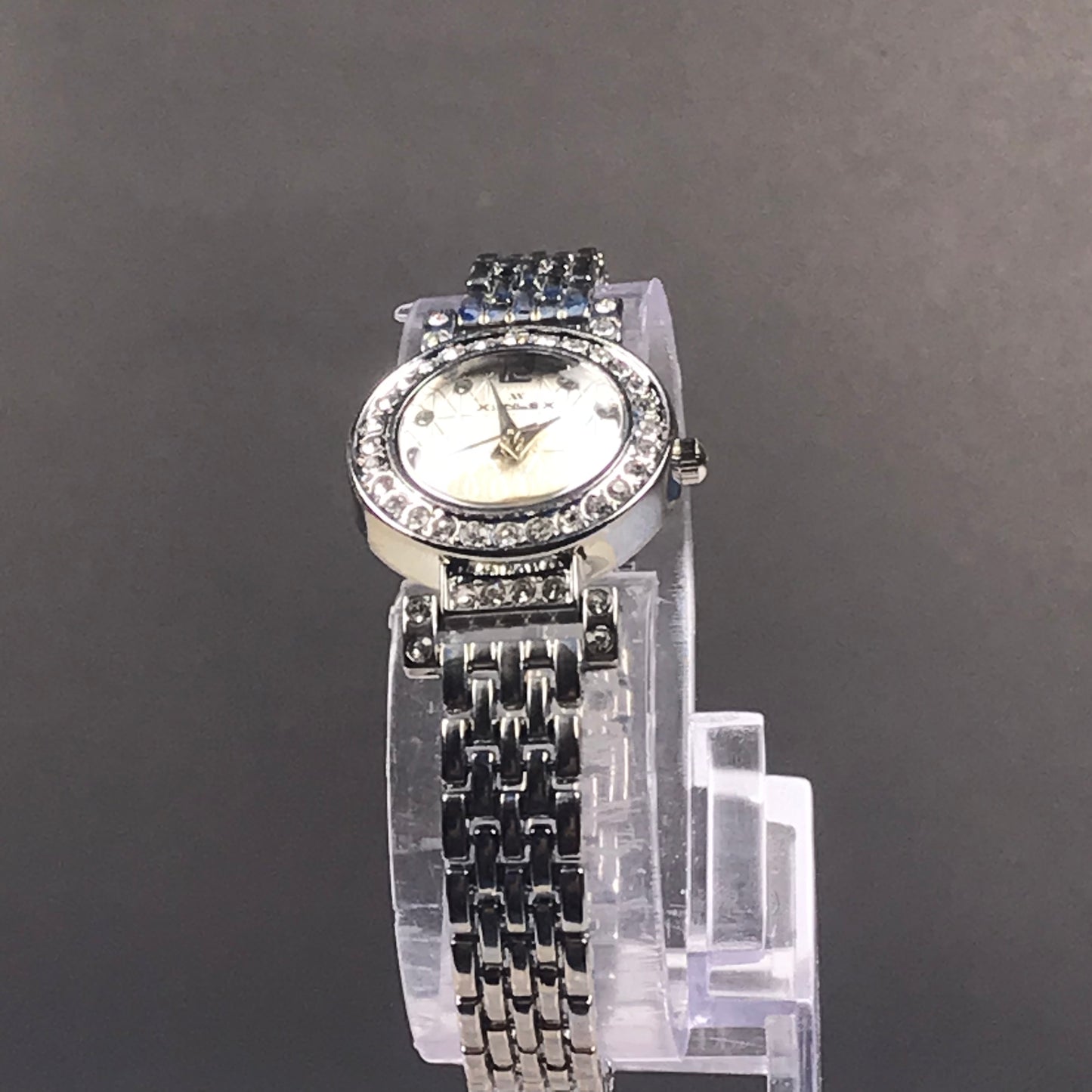 A women's silver-tone wristwatch with a rhinestone-studded bezel, white dial, and matching rhinestone-embellished bracelet, displayed on a clear stand against a dark gray background.