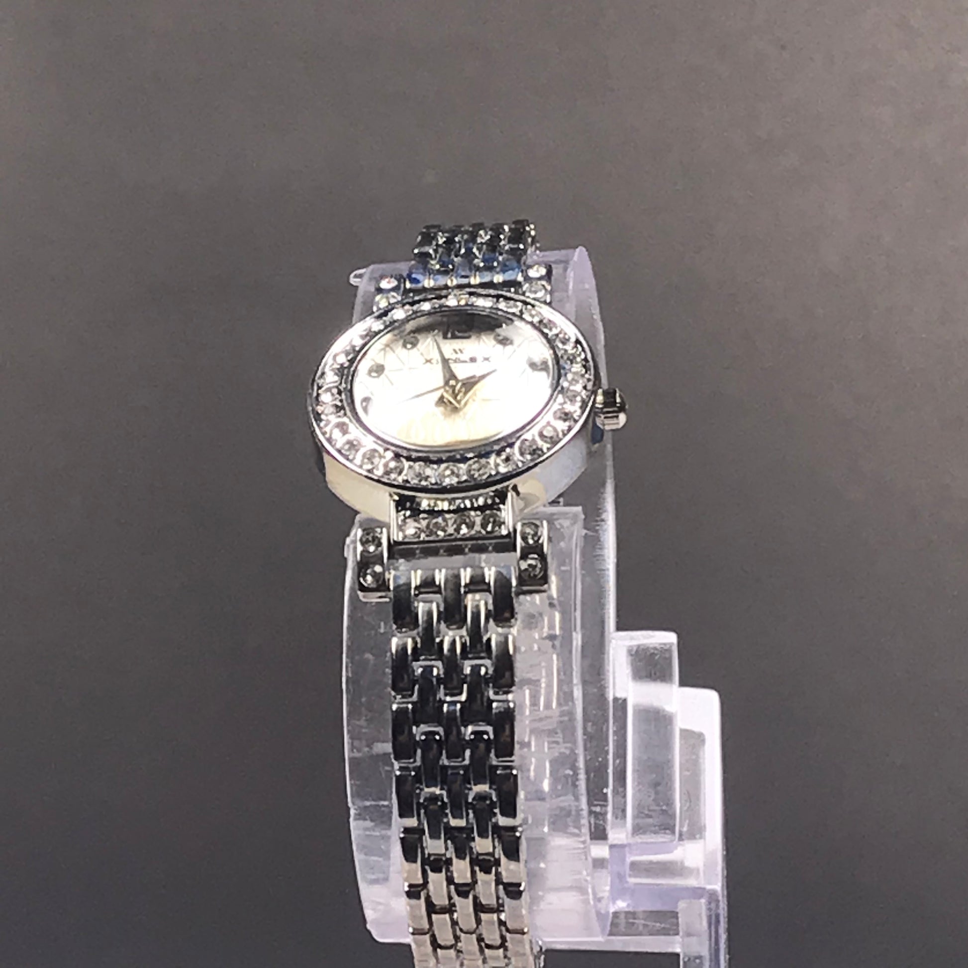 A women's silver-tone wristwatch with a rhinestone-studded bezel, white dial, and matching rhinestone-embellished bracelet, displayed on a clear stand against a dark gray background.