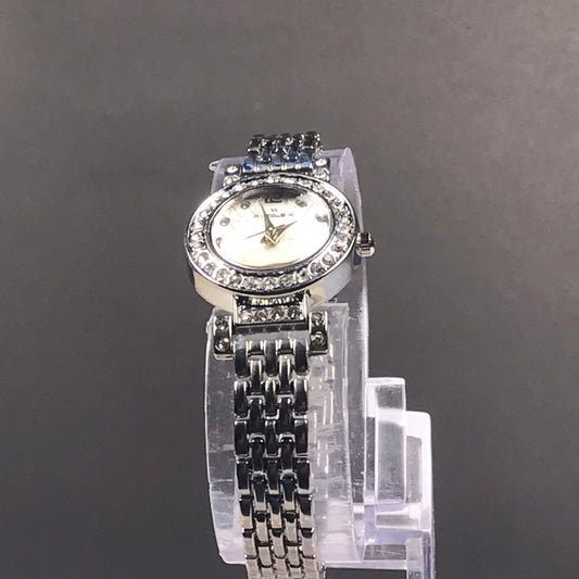 A women's silver-tone wristwatch with a rhinestone-studded bezel, white dial, and matching rhinestone-embellished bracelet, displayed on a clear stand against a dark gray background.