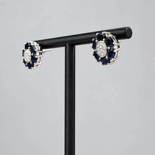 A pair of round cluster stud earrings with dark blue stones and a central clear stone, set in silver-toned metal, displayed on a black T-stand against a light gray background.