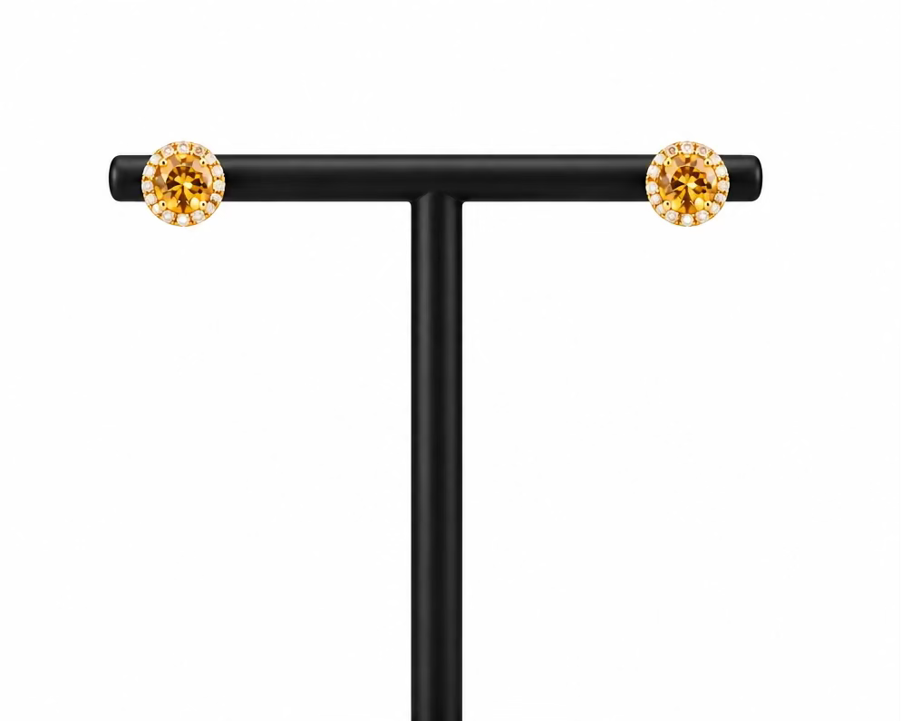 A pair of round stud earrings with sparkling clear stones in a gold-toned bezel setting, displayed on a black T-stand in front of a white Meenu Jewels box against a dark gray background.