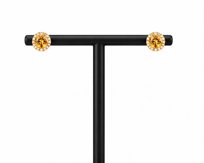 A pair of round stud earrings with sparkling clear stones in a gold-toned bezel setting, displayed on a black T-stand in front of a white Meenu Jewels box against a dark gray background.