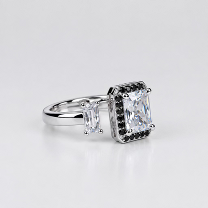 A silver-toned ring with a large rectangular clear gemstone and smaller side stones, displayed on a light surface with a blurred white jewelry box in the background.