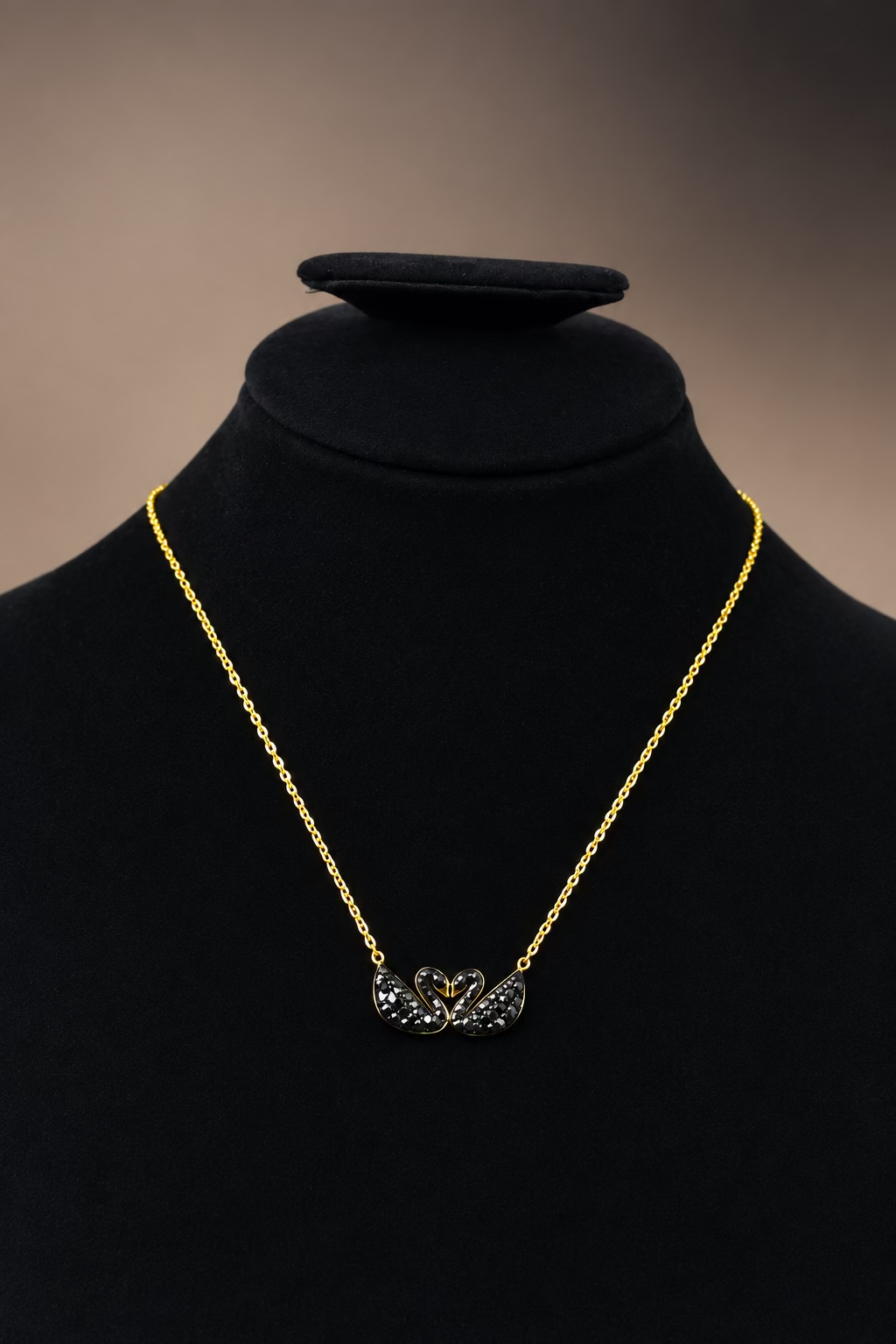 A delicate gold chain necklace with a small black crystal-encrusted pendant, displayed on a black velvet jewelry bust against a dark gray background.