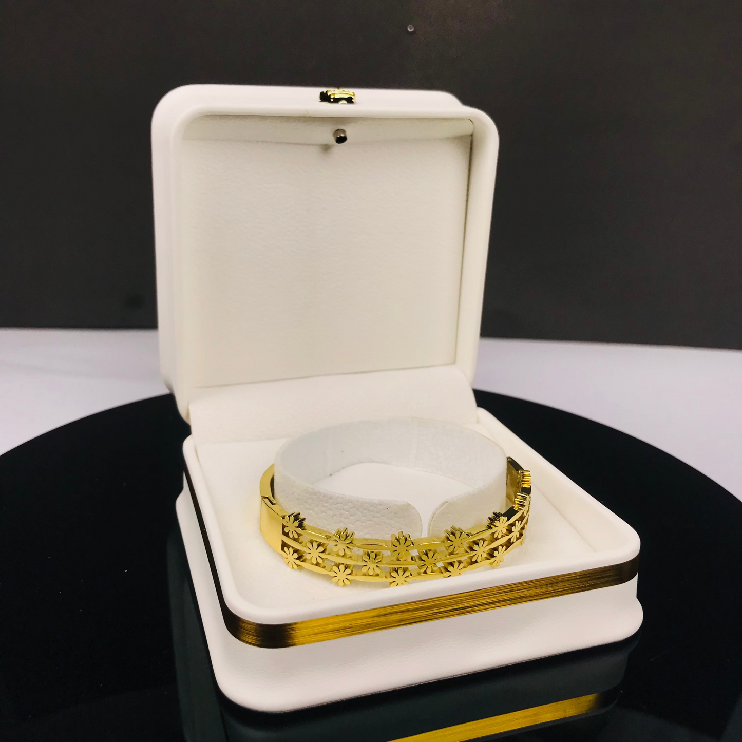 A gold-toned ornate bracelet displayed inside an open white jewelry box with gold trim, set on a reflective black surface against a dark gray background.