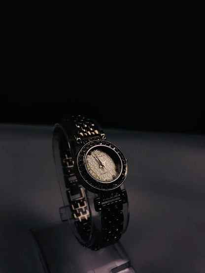 A black women's watch with a textured dial and a black link bracelet accented with gold-tone details, displayed on a stand against a dark background.