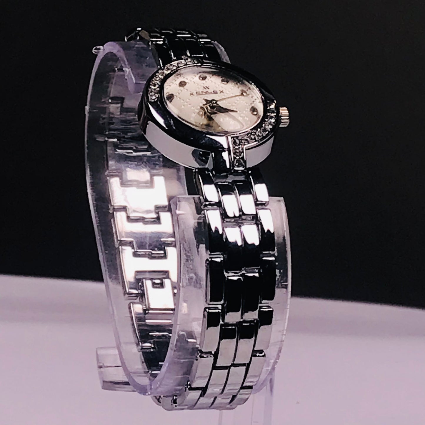 A women's analog watch with a silver-tone metal link bracelet, a black bezel, and a white dial with crystal accents, displayed on a clear stand against a black and white background.