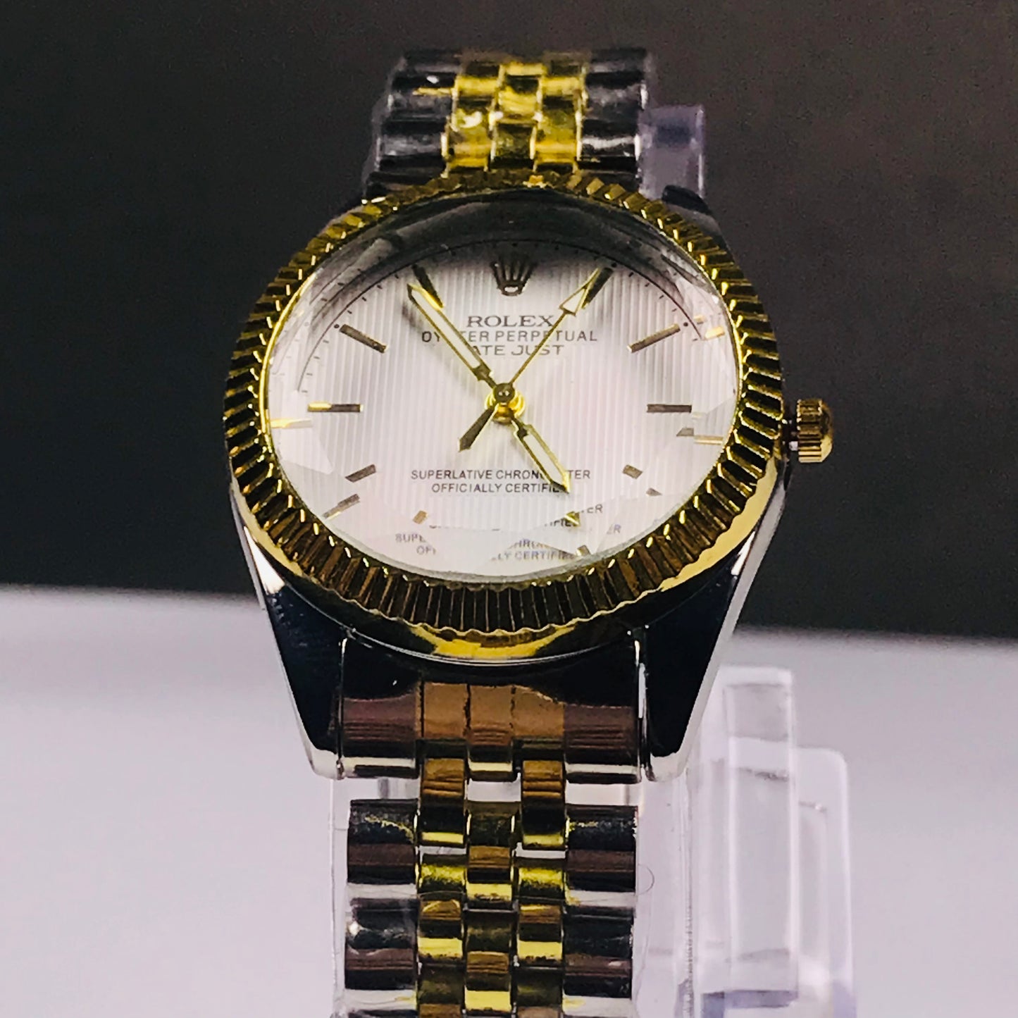 A Rolex wristwatch with a two-tone yellow gold and stainless steel link bracelet, a fluted gold bezel, and a white dial with gold hands and markers, displayed on a clear stand.