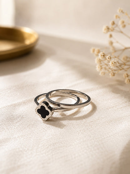 Two silver rings on a textured cream fabric background, with one ring featuring a black clover-shaped stone surrounded by a small silver border.
